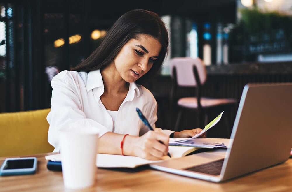 Fundo de uma cafeteria. Mulher de cabelos longos e escuros, sentada em uma mesa segurando papéis e escrevendo em um caderno na frente de um notebook, com um copo de café ao seu lado.
