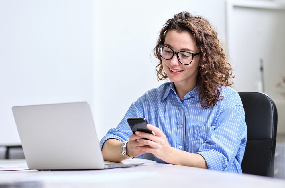 Fundo de uma sala. Mulher de óculos e camisa azul sentada em uma mesa olhando para um celular em frente a um notebook.